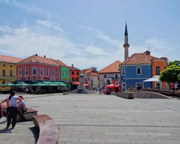 Kolorowy plac w **Tuzla, Bośnia**, z kawiarniami, fontanną i minaretem w tle.