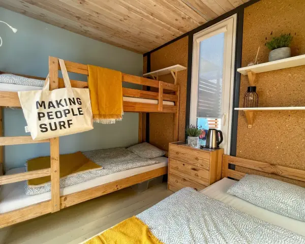 Wnętrze chałupy kempingowej z łóżkiem piętrowym i pojedynczym, żółtym kocem i torbą z napisem "MAKING PEOPLE SURF".
