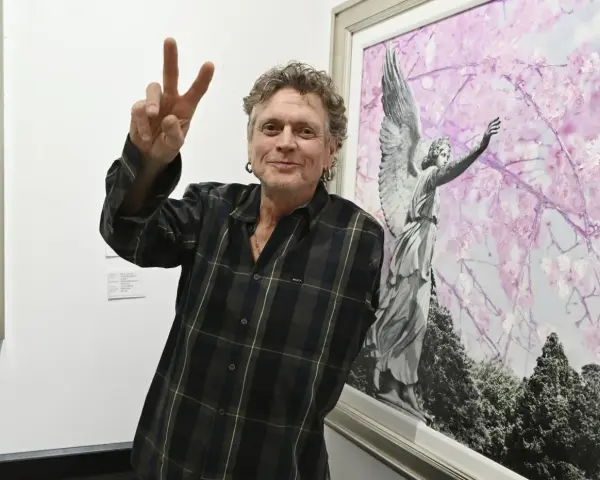 Rick Allen, perkusista Def Leppard, pozuje z uniesionym znakiem pokoju obok obrazu z aniołem i kwitnącymi drzewami.