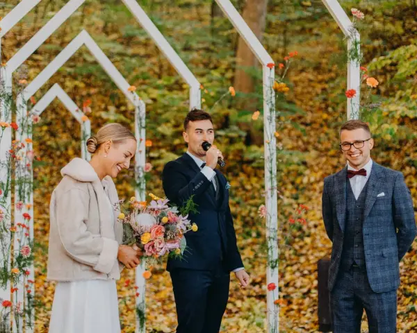 Uroczyste wesele w listopadzie. Panna młoda z bukietem, pan młody przemawia do mikrofonu, a świadek uśmiecha się w tle.