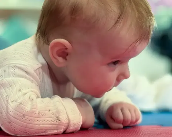 Malutkie dziecko leży na brzuchu, z zaciekawieniem spoglądając przed siebie. Jego duża głowa u dziecka jest lekko pochylona, a drobne rączki podparte o podłoże.