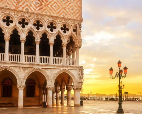 Palazzi storici di Venezia all'alba, con il sole che illumina la piazza e le architetture gotiche.