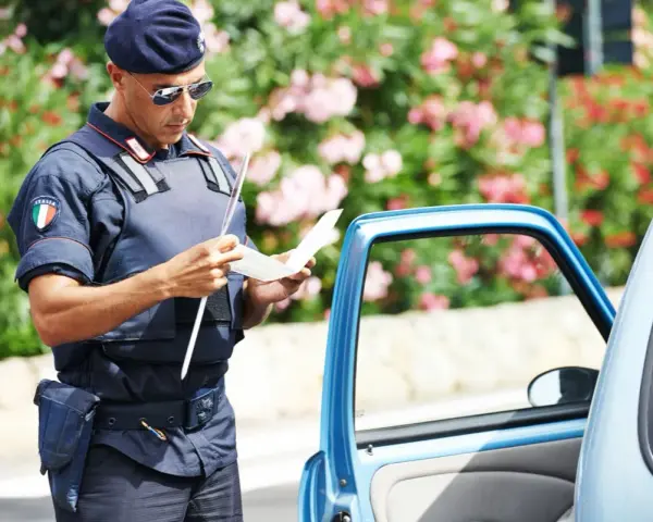 Agente di polizia italiana con giubbotto antiproiettile e berretto blu, controlla un verbale vicino a un'auto blu. Potrebbe essere un controllo legato all'art. 180 cds.