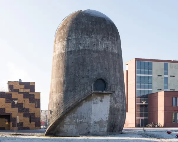 Der Trudelturm Berlin, ein futuristisches Betonbauwerk, steht neben modernen Gebäuden. Eine Außentreppe führt zu einer runden Tür.