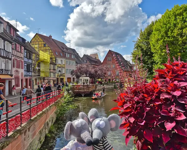 Deux peluches souris posent devant les maisons colorées de Colmar, une destination idéale pour visiter Colmar en 1 jour. Une barque navigue sur le canal.
