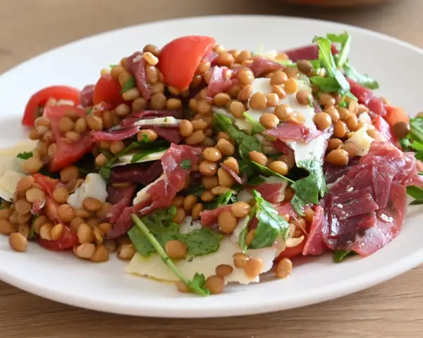Insalata di lenticchie con pomodorini, scaglie di parmigiano e prosciutto crudo, un piatto fresco e gustoso.