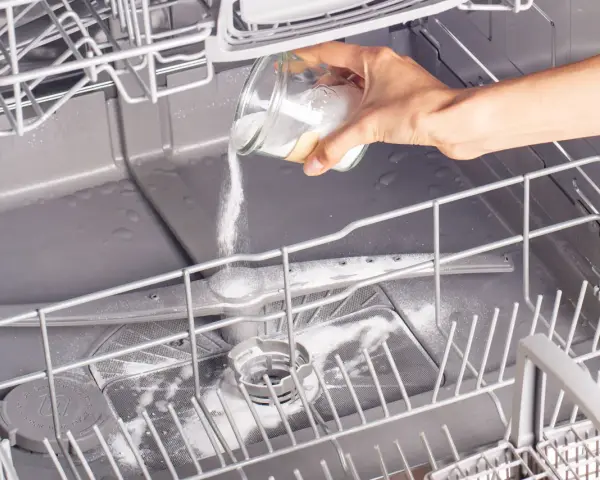A hand pours white powder from a jar into an open dishwasher, demonstrating how to clean a dishwasher naturally.