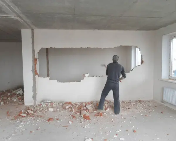 Mężczyzna wyburza młotem duży fragment ściany nośnej, tworząc otwór. Wokół leżą gruzy.