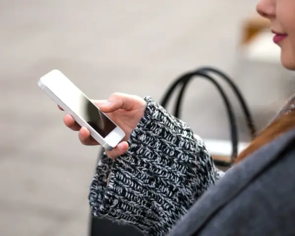 Mano che tiene uno smartphone, con un maglione a trecce e una borsa sullo sfondo. La persona sta utilizzando dati per navigare.