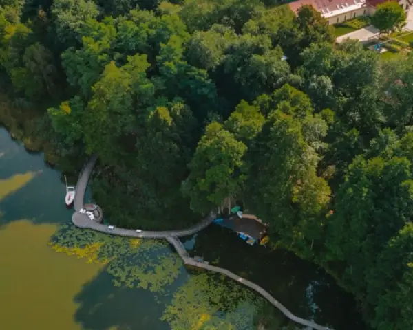 Żaglówka i deska SUP na spokojnym jeziorze ostrowskim, otoczonym bujną zielenią i drewnianą kładką.