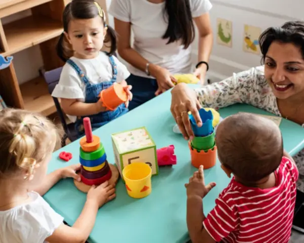 Zwei Frauen und drei Kleinkinder spielen mit bunten Spielsachen. Eine Frau hilft einem Kind, Türme zu stapeln, was eine tolle Methode ist, um Kinder zweisprachig zu erziehen.