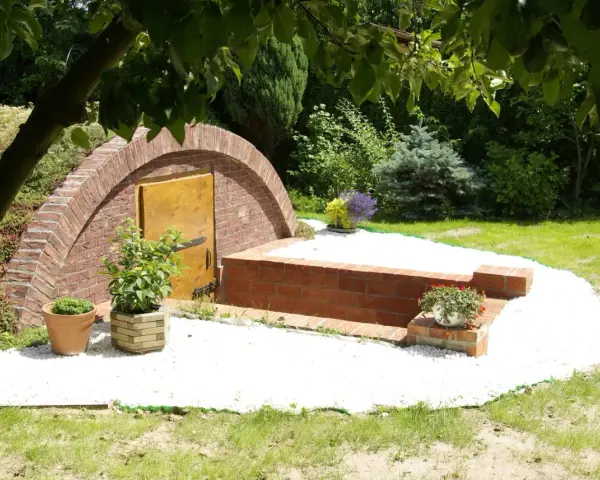 Urokliwa piwnica w ziemi, zbudowana z cegły, z drewnianymi drzwiami i otoczona białym żwirem. Wokół kwitnące rośliny i zieleń.