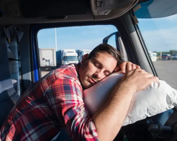 Ein LKW-Fahrer schläft auf einem Kissen im Führerhaus. Das Bild thematisiert die Einhaltung des Arbeitszeitgesetzes für LKW-Fahrer.