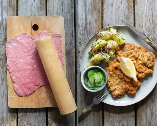 Förberedelser för schnitzel recept: tunt bankat kött på skärbräda, bredvid en tallrik med schnitzel, potatissallad och gurka.