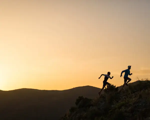 Dos corredores se enfrentan a una exigente carrera de senderos al amanecer, sus siluetas recortadas contra un cielo anaranjado.
