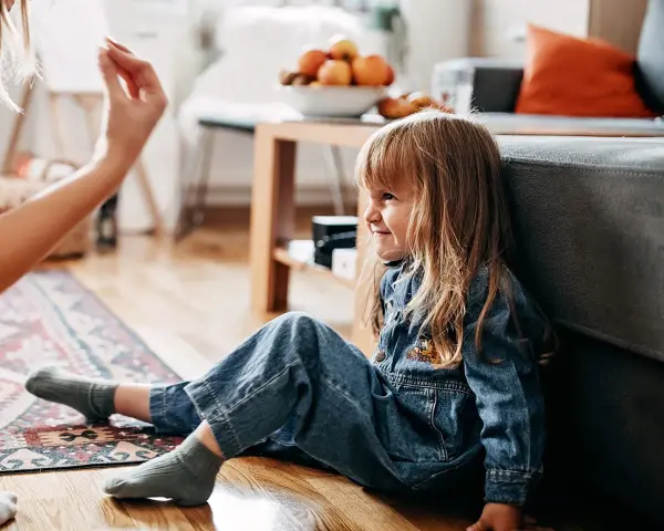Dziewczynka w dżinsowym ogrodniczku siedzi na podłodze, uśmiecha się. Mama wyciąga rękę. Brak oznak agresji u dzieci.