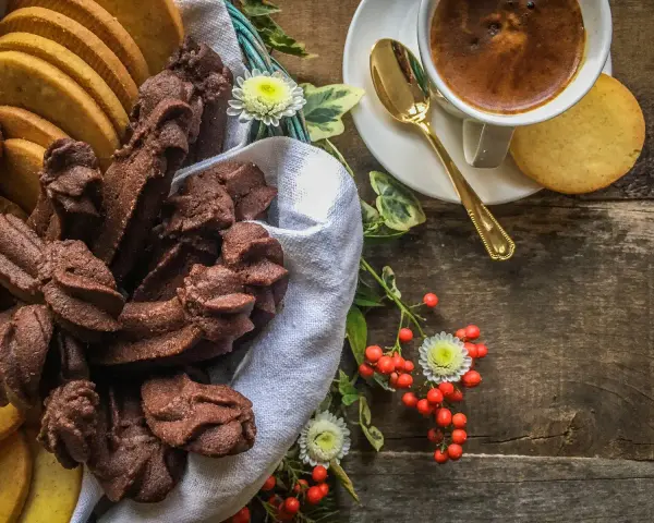 Cesto pieno di biscotti fatti in casa al cioccolato e alla vaniglia, accanto a una tazzina di caffè e bacche rosse.