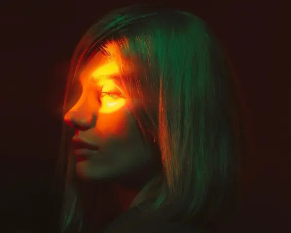 Profilo di donna con luce arancione sull'occhio, evocando un'emicrania oculare. Capelli illuminati da luci verdi e rosse.
