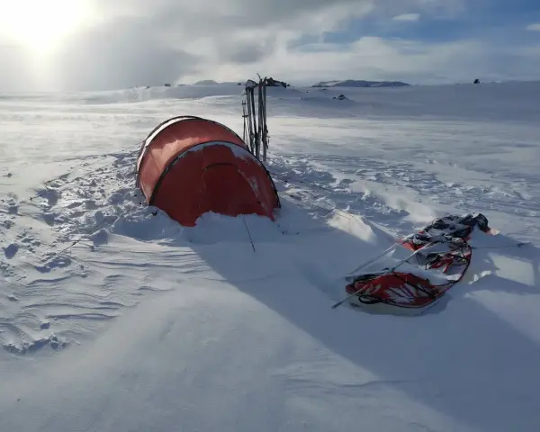 Ein rotes Wintercamping Zelt steht im Schnee, daneben Skier und ein Schlitten. Die Landschaft ist weit und verschneit.