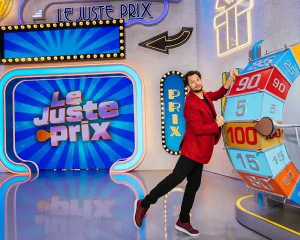 Un animateur pose près de la roue du jeu "Le Juste Prix". L'illustration du plateau est colorée et lumineuse, promettant de beaux gains.