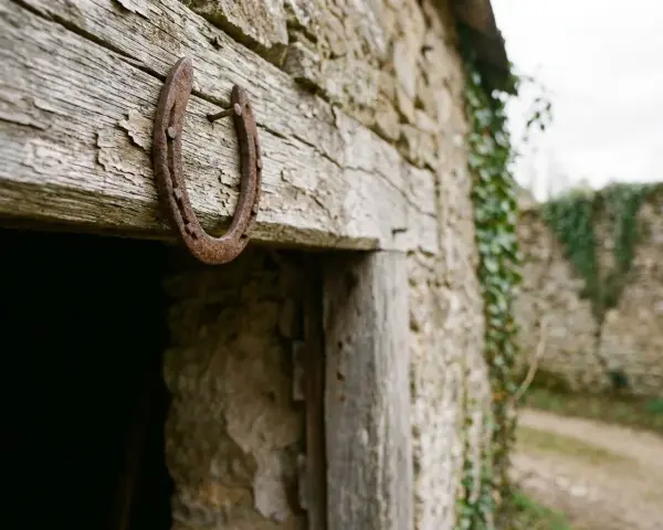 Un vieux fer à cheval pointes en bas, rouillé, est cloué sur une poutre en bois patiné, devant une bâtisse en pierre recouverte de lierre.
