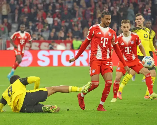 Intensywna akcja na boisku podczas meczu składy: Borussia Dortmund – Bayern Monachium. Piłkarz Bayernu w czerwonej koszulce próbuje odebrać piłkę leżącemu zawodnikowi BVB.