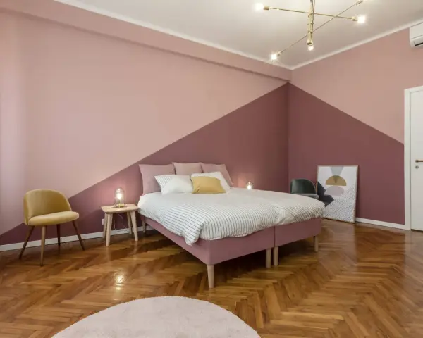 Camera da letto con pareti bicolore, un letto rosa, una sedia gialla e un parquet a spina di pesce. Un esempio di come pitturare una stanza con stile.