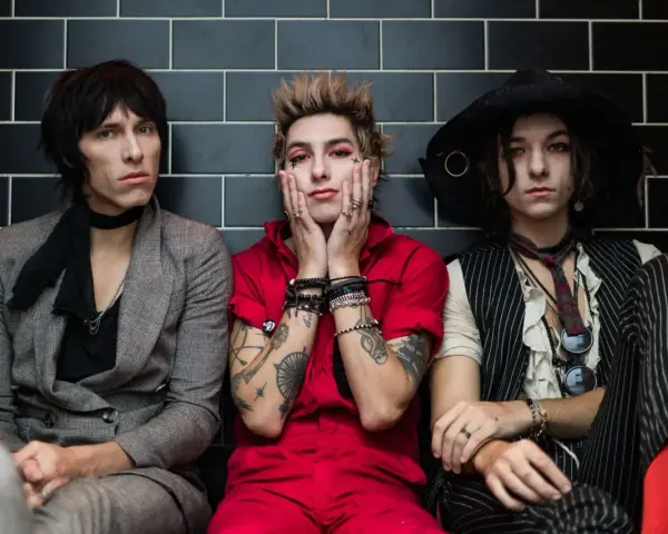 Trzech członków Palaye Royale w stylowych, rockowych strojach siedzi obok siebie na tle czarnych płytek.