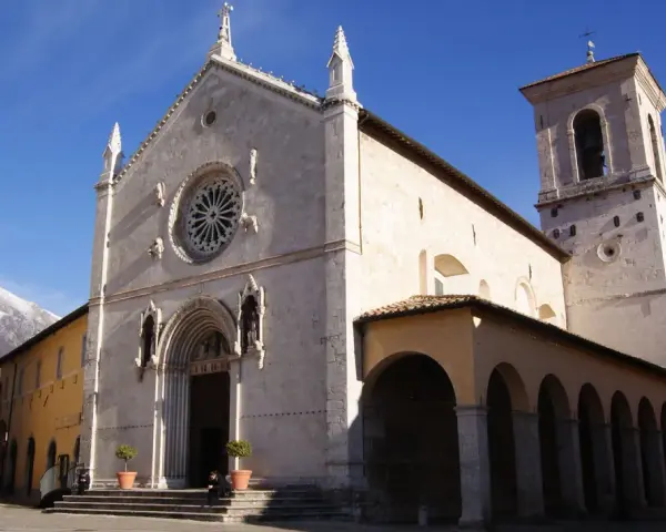 A grand church with a rose window and bell tower stands in a town square, with snow-capped mountains in the distance. This is Nursia, a historic Italian town.