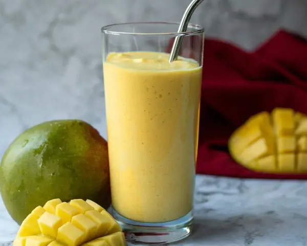 Orzeźwiający koktajl z mango w szklance, obok świeże mango i kawałki owocu. Idealny na upalne dni.