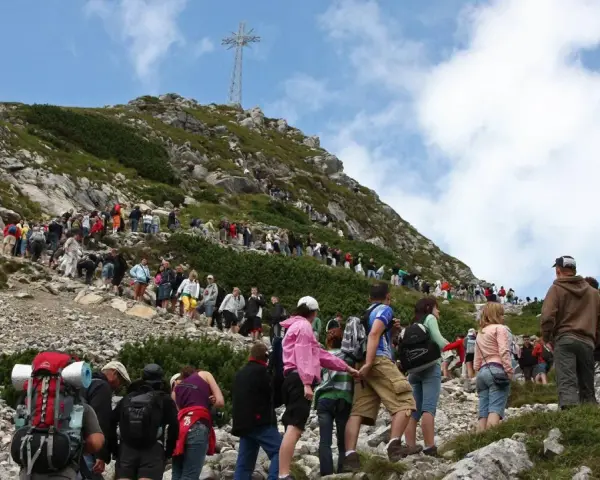 Tłum turystów wspina się na Giewont, podziwiając widok z Kasprowego Wierchu. Na szczycie widoczny krzyż.