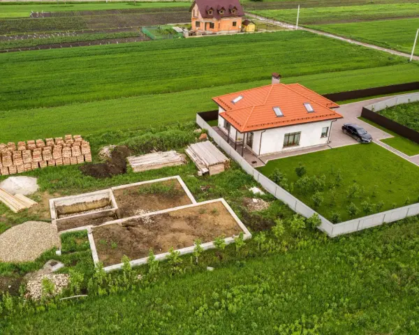 Działka 1000m2 wymiary: rozpoczęta budowa domu, gotowy fundament, sterta cegieł, drewno, żwir, obok stoi dom z pomarańczowym dachem i samochód.