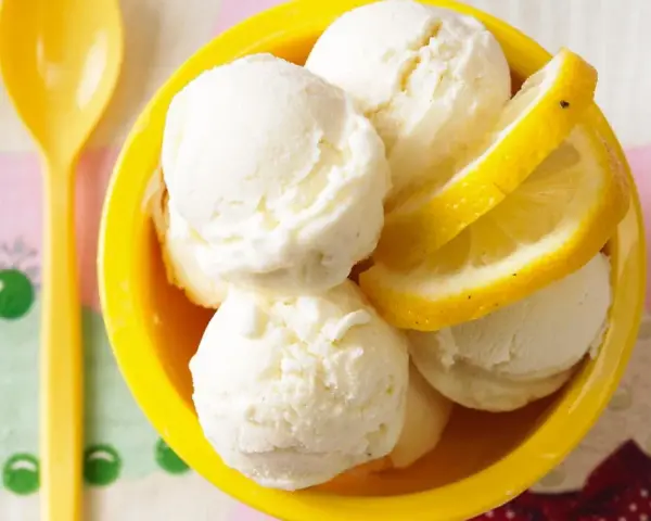 Erfrischendes Buttermilch Zitronen Eis in einer gelben Schale, garniert mit Zitronenscheiben. Ein gelber Löffel liegt daneben.
