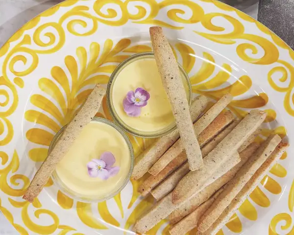 Två skålar med gul kräm, dekorerade med violer, serveras med krispiga kex på en gulmönstrad tallrik. En perfekt påsk dessert.