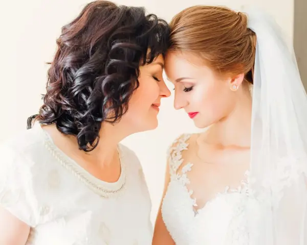Matka i córka, obie elegancko na ślub córki, dzielą wzruszający moment. Panna młoda w białej sukni, mama w ozdobnej bluzce.
