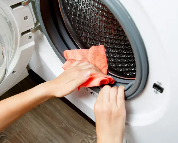 Hands cleaning the rubber seal of a washing machine with an orange cloth. This is part of how to clean washing machine.