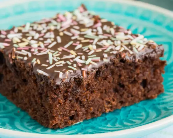 Ein Stück saftiger Schokoladen-Blechkuchen mit Schokoladenglasur und bunten Streuseln auf einem türkisfarbenen Teller. Ein einfaches Blechkuchen Rezept für Glücksmomente.