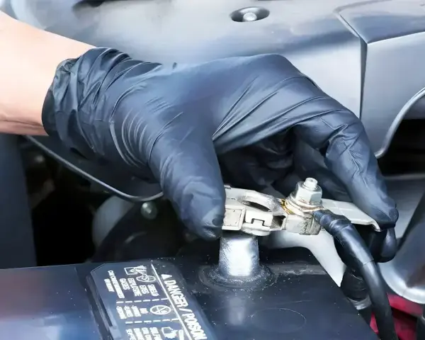 Gloved hand applying WD40 to a car battery terminal to prevent corrosion and ensure a good connection.