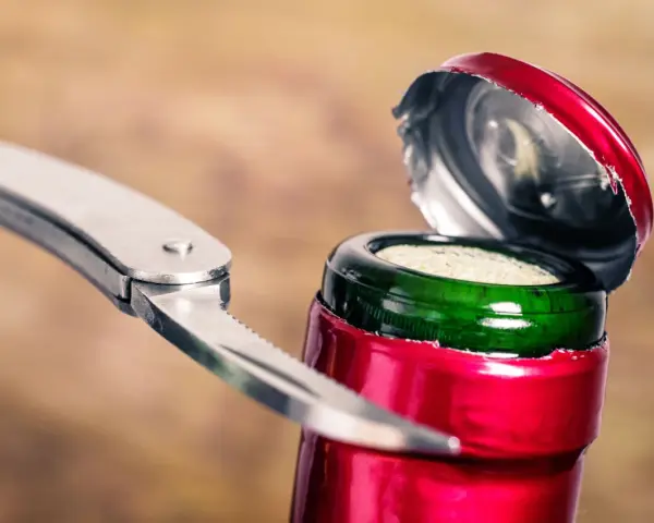 Metalowy widelec próbuje otworzyć butelkę wina, podważając czerwone wieczko.