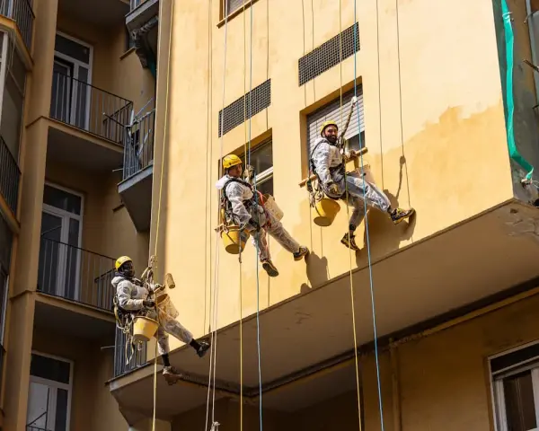 Operai in corda applicano diversi tipi di intonaco per esterni su un edificio giallo.