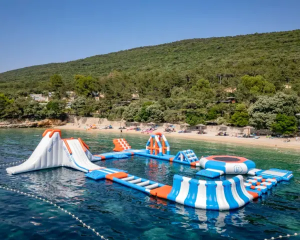 Wodne atrakcje na krystalicznie czystej wodzie, idealne na wakacje w **camping Slatina**. W tle zielone wzgórza i plaża.