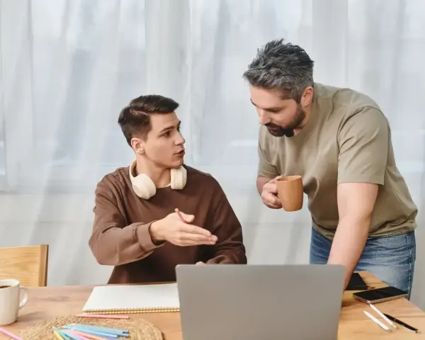 Ojciec rozmawia z synem, pokazując mu, jak zmotywować dziecko do nauki. Na stole laptop, zeszyt i kubek kawy.
