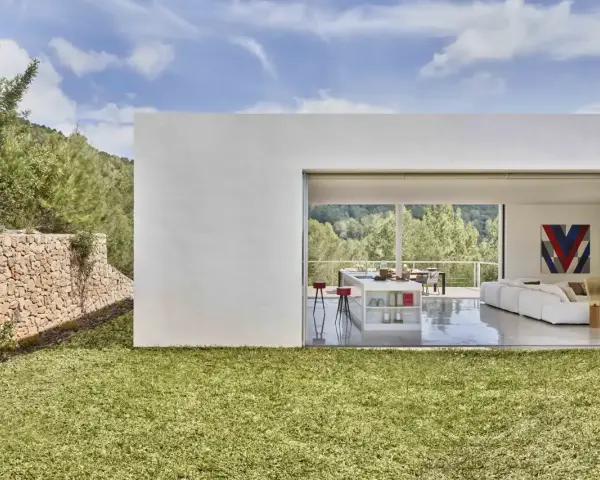 Exterior de casa ibicenca con césped verde y muro de piedra. Interior moderno con cocina y salón abiertos al paisaje.
