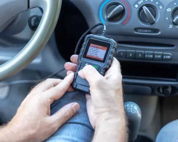 Ręce trzymające skaner diagnostyczny, który pokazuje menu z opcją "02 Sensor Test". Urządzenie może pomóc zdiagnozować problemy z czujnikiem spalania stukowego.