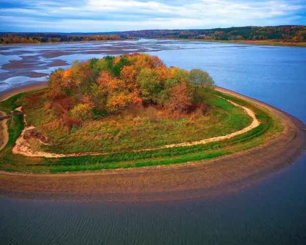 Jesienna wyspa na zalewie Brody, porośnięta kolorowymi drzewami i trawami, otoczona błękitną wodą.