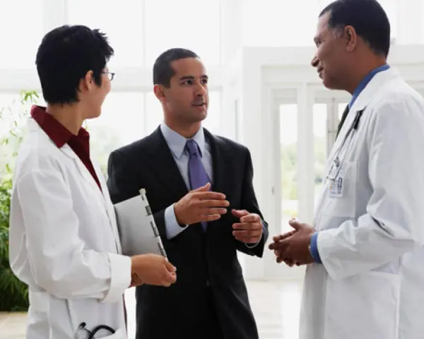 Un uomo in giacca e cravatta parla con due medici in camice bianco. Forse discutono di un nuovo farmaco e del suo informatore scientifico stipendio.