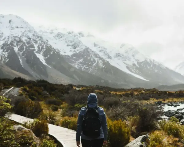 Une personne avec un sac à dos fait une randonnée sous la pluie, face à des montagnes enneigées. Le chemin en bois serpente à travers une végétation basse.