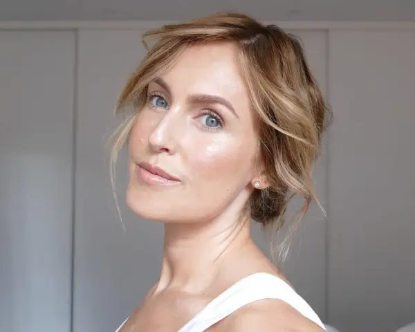 En kvinna med blont hår och blå ögon har ett naturligt glow i ansiktet. Hon bär en vit topp och en liten diamantörhänge.
