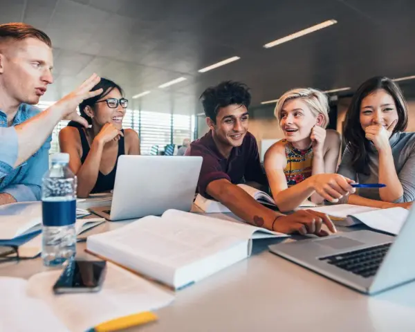 Fünf Studierende lernen gemeinsam. Sie lachen und diskutieren, während sie an Laptops arbeiten. Ein Mann gestikuliert lebhaft, was auf ein spannendes Studium im Ausland hindeutet.