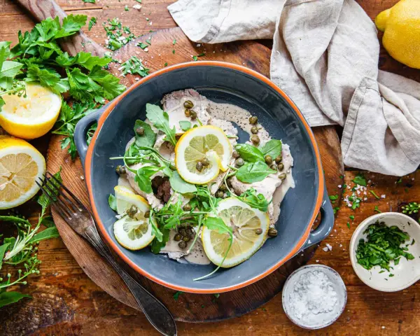 Vitello tonnato i en skål, toppad med citronskivor, kapris och ruccola. Serveras med citroner och färska örter.